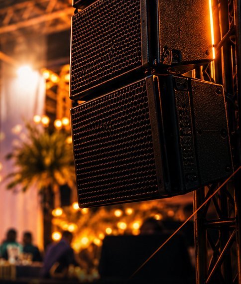 Caixas de som em evento com iluminação quente e convidados ao fundo